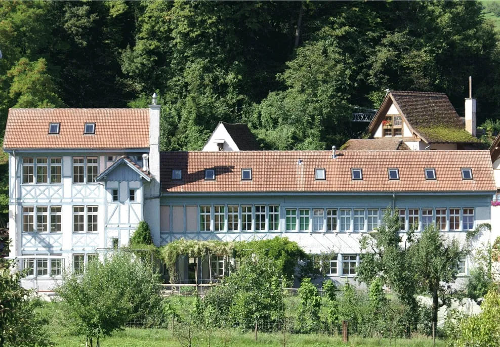 Die Alte Fabrik in Maisprach: ein weiss verputzter Altbau mit Sprossenfenstern und ziegelrotem Dach, dahinter ein Haus mit Holzgiebel und Wald.