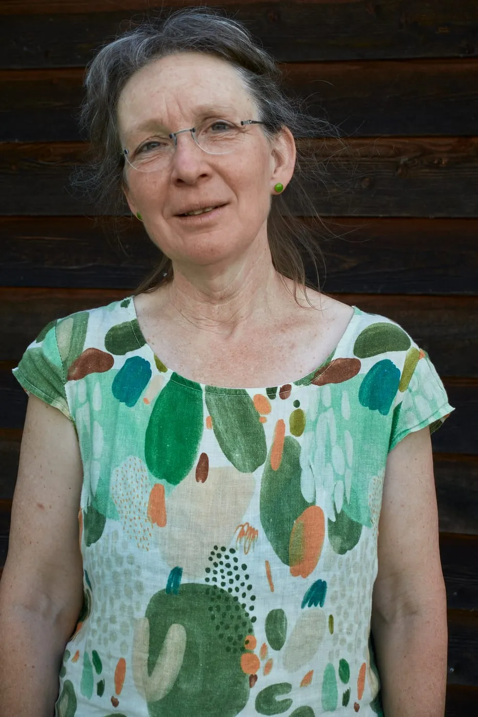 Monika Sennhauser, frontal vor dunkler Holzfassade, in einem weissen Oberteil mit grünem Blattmuster.